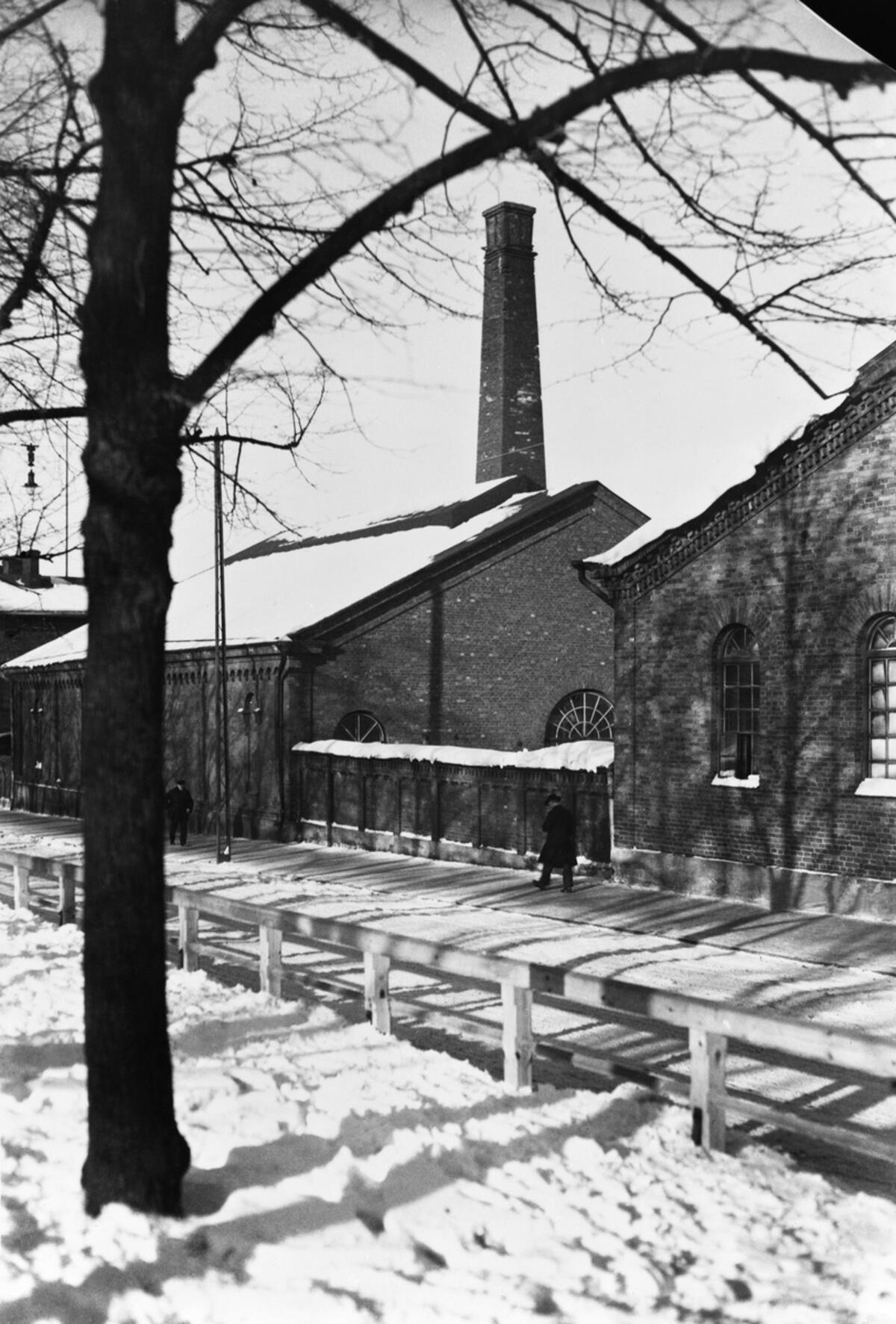 Bilden som tagits en vintrig dag visar fotgängare, bakom dem ligger tegelbyggnader av ett gasverk. Bilden har tagits från en parkgång som gick i mitten av nuvarande Mannerheimvägen år 1930.