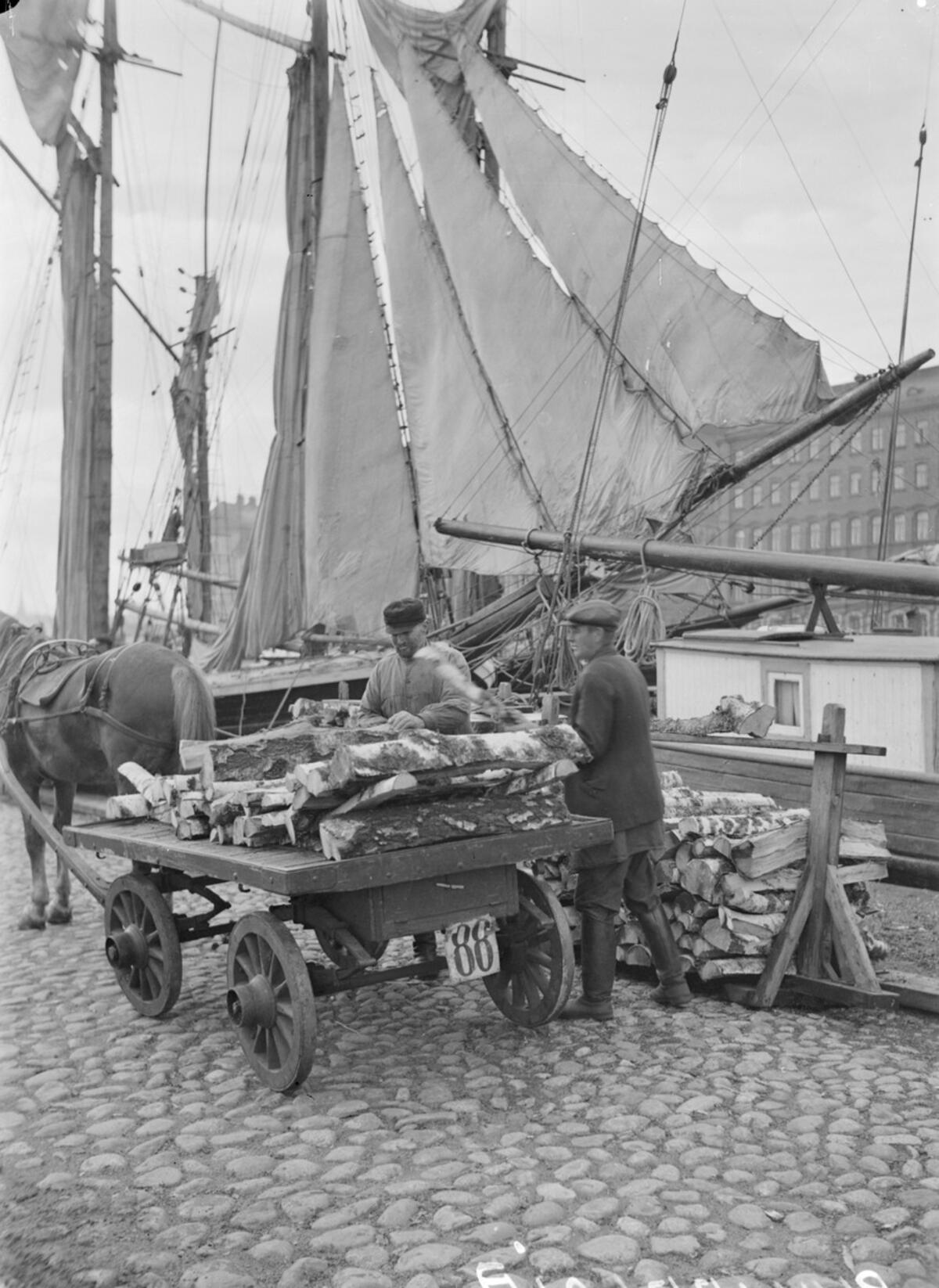 Men stacking firewood in a horse-drawn cart at the Halkolaituri pier in the North Harbour of Kruununhaka; yawls loaded with wood can be seen in the background.