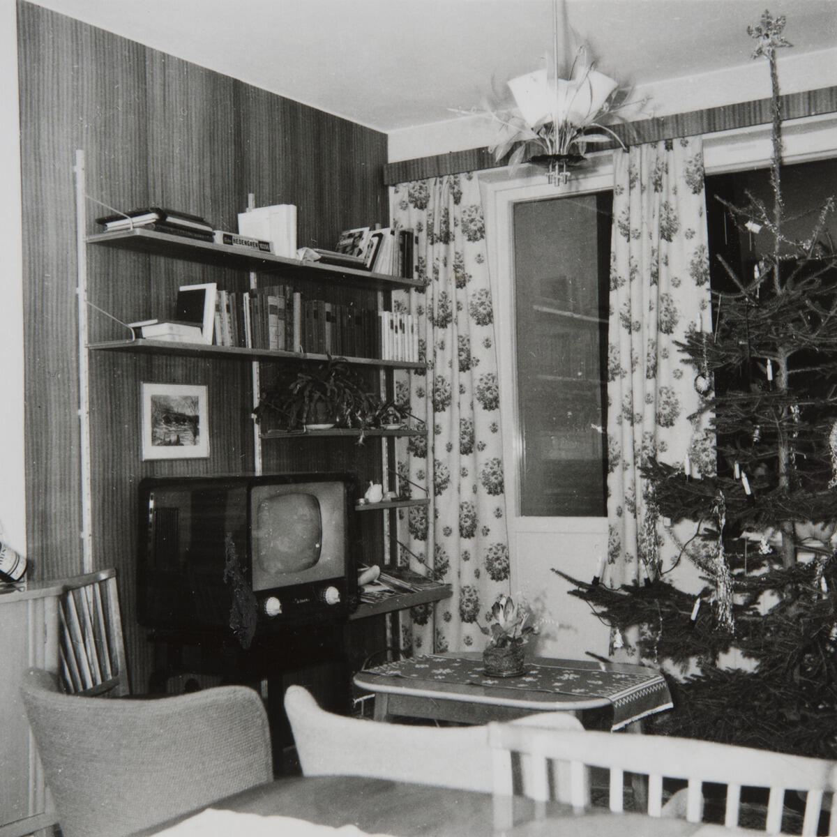 Till vänster en bokhylla med tv, till höger en julgran. I förgrunden stolar och ett bord.