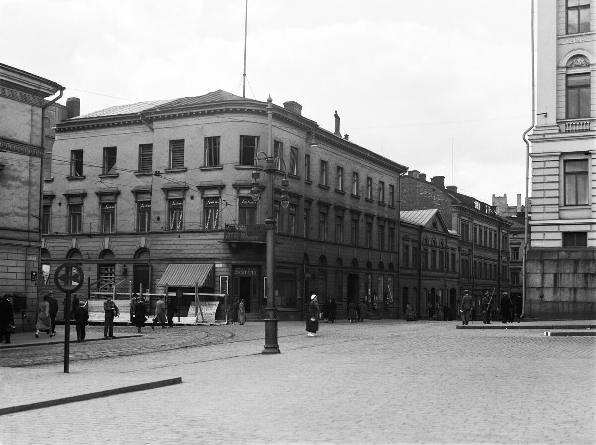 Hörnhuset har tre våningar. I mitten av bilden står en gatlykta. I vänstra kanten skymtar Kiseleffska huset, i högra universitetets huvudbyggnad.