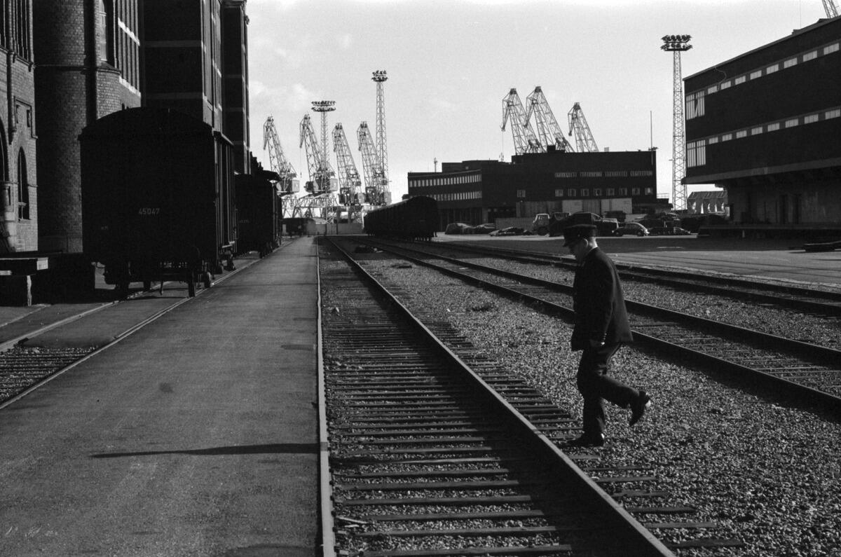 Utsikt mot sydost från Skatuddskajen. I mitten av bilden löper fyra par tågskenor, och en man håller på att korsa bangården. Till vänster ett godståg. På båda sidorna av skenorna står magasinsbyggnader, och i bakgrunden flera lyftkranar.