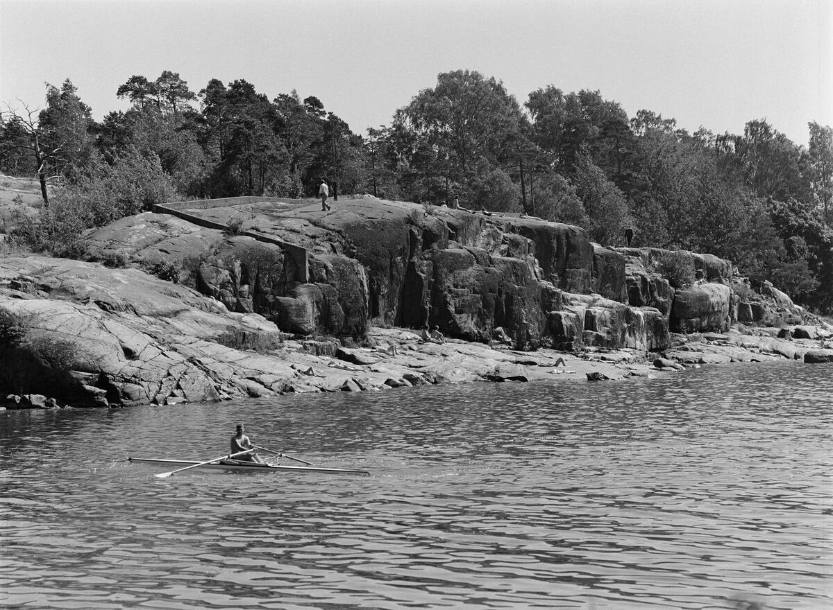 Auringonottajia ja soutava mies Humallahden rannassa, Meilahdessa. Kallioilla näkyy vielä jo pois puretun Humallahden uimalaitoksen perustuksia.