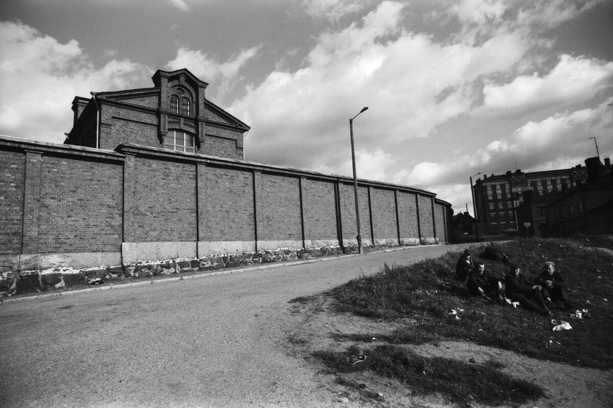 On the left is a prison behind a brick wall. On the right a group of men sitting on the grass drinking alcohol.
