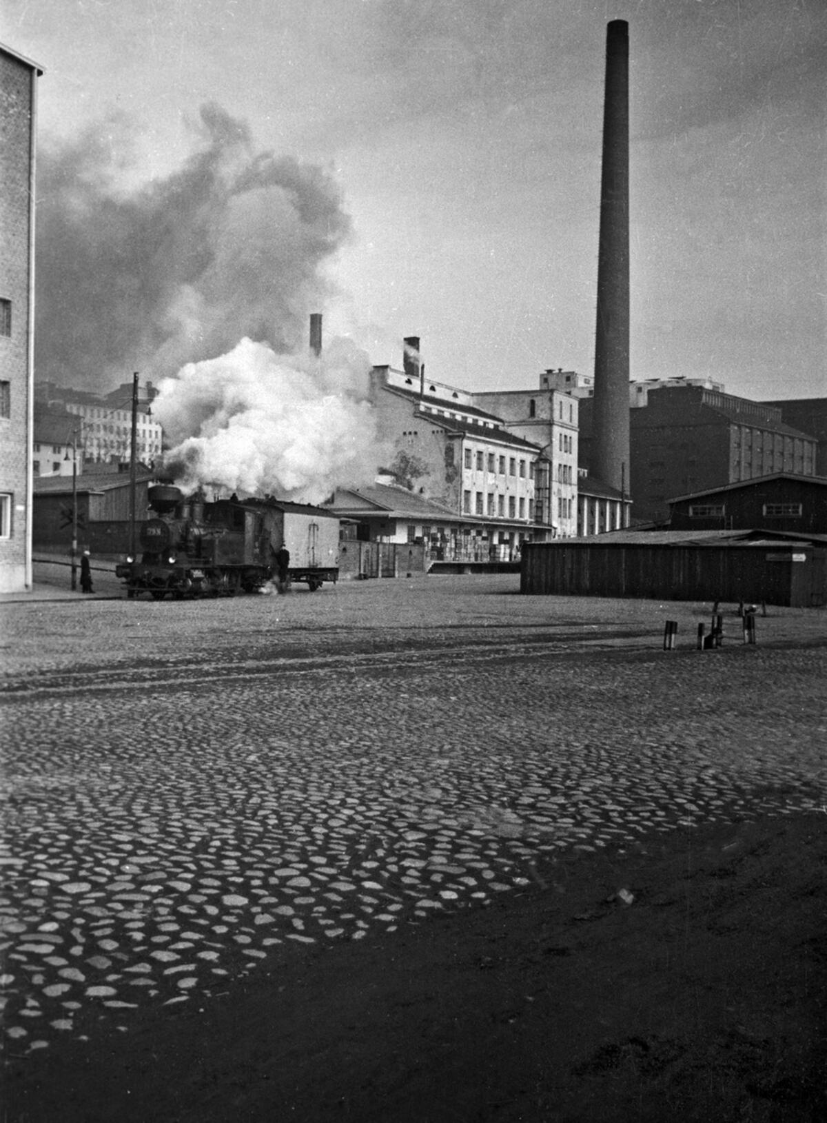 "Pässi" eli päivystävä veturi Lintulahdenkujan ja Kaikukadun risteyksessä. Päivystävä veturi piti huolen siitä, että tyhjiä junavaunuja oli siellä missä niitä tarvittiin Sörnäisten satamassa. Höyryveturin piipusta nousee savua, taustalla on tehdasrakennuksia.