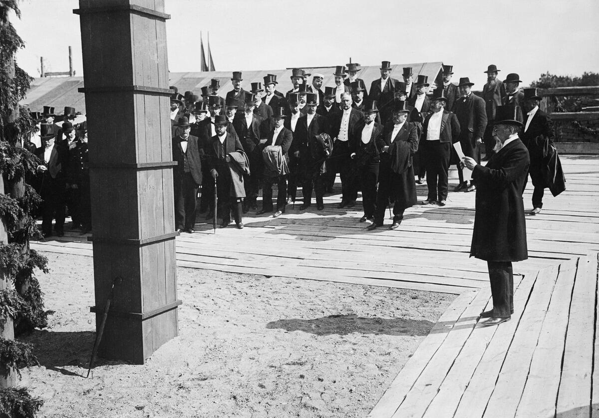 En man som är klädd i cylinderhatt och knälång rock håller ett tal med sidan mot publiken. I bakgrunden står en grupp festklädda män, av vilka en del har frack och cylinderhatt.