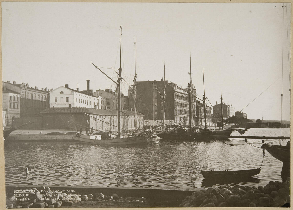 Träbåtar i hamnen. Vid stranden står låga magasinsbyggnader och i bakgrunden höghus av sten.