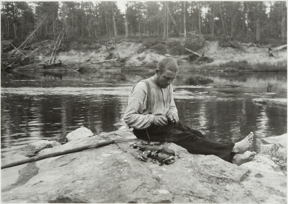 Mies istuu kalliolla veden ääressä ja avaa veitsellä raakkua. Hänen vieressään on kasa raakkuja.