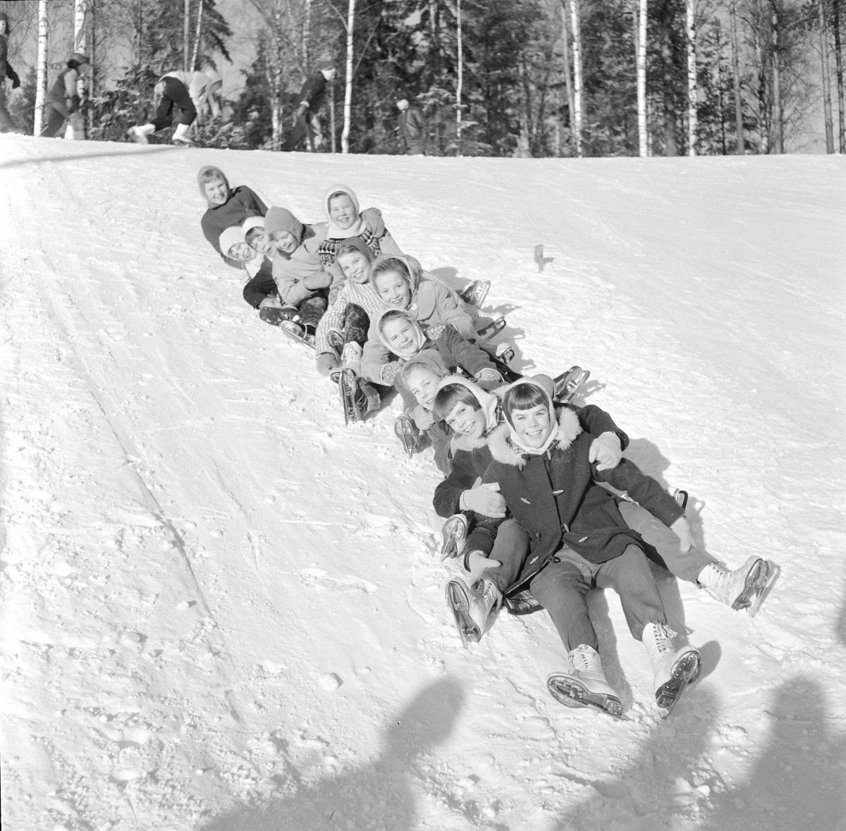 Yli kymmenen lasta on muodostanut pitkän letkan, joka laskee yhdessä pyllymäkeä. Ainakin etummaisilla on luistimet jalassa. 