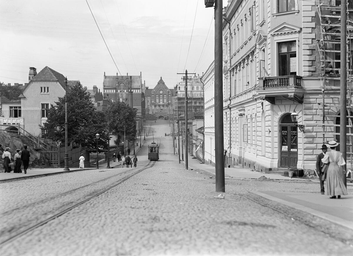 A view of Nikolainkatu (now Snellmaninkatu) and the buildings along it. At the end of the street, you can see the University of Helsinki Department of Physiology (now the Faculty of Behavioural Science). There is only one set of tram tracks with a tram on them. There is also a horse-drawn carriage and people walking on the pavement.