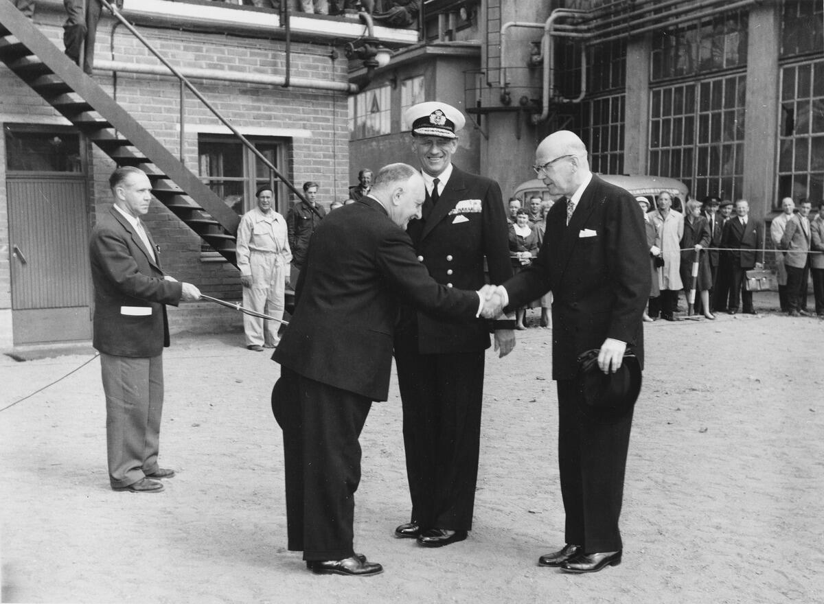 Längst fram i mitten skakar bergsrådet Wahlforss och president Kekkonen hand med sidan vänd mot tittaren. Bakom dem står den leende kungen av Danmark iförd flottans hatt. I bakgrunden syns varvsbyggnadens ytterväggar och bakom ett rep står åskådare, varav en del är klädda i arbetsoveraller.