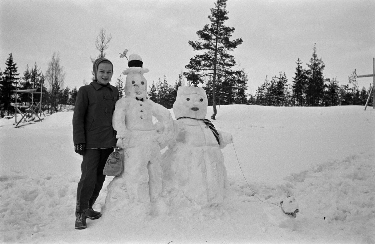 Ett barn med sina snöalster: två snögubbar och en snökatt eller -hund. Snögubbarna föreställer Kalle Träskalle och hans fru.