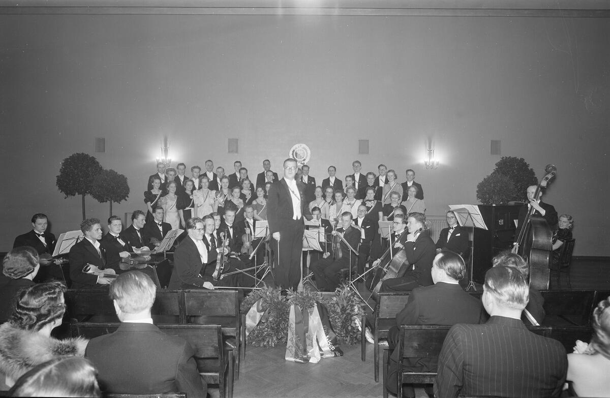 Mitt på bilden syns en orkester och en kör. I förgrunden syns publik med ryggen mot kameran.