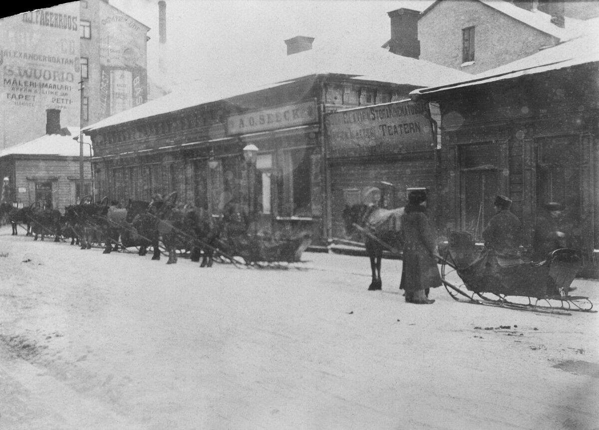  Hyrkuskar med sina hästdrivna slädar på Glogatan 3 på vintern i början av 1900-talet. Bakom dem står låga trähus.