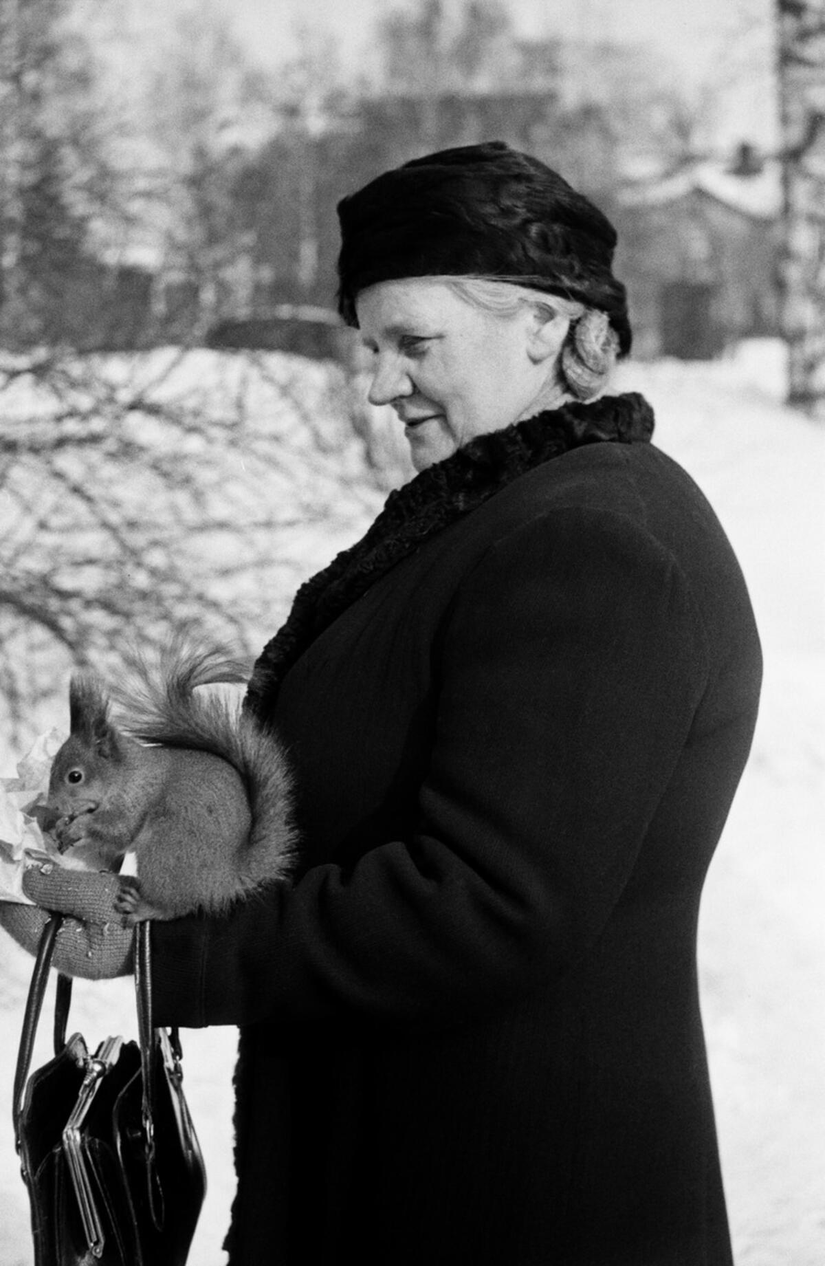 Mustavalkoinen valokuva, jossa nainen on sivuttain kameraan. Hänen kädellään istuu orava syömässä. Nainen hymyilee, taustalla on lunta.