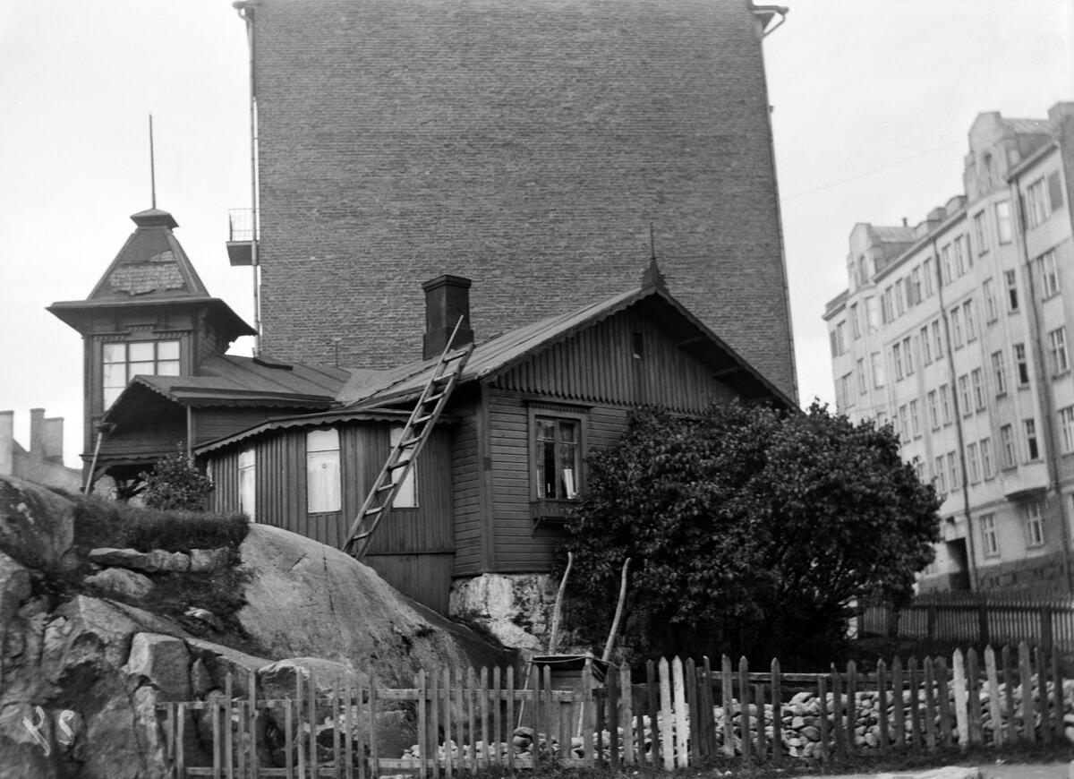 Ett lågt tröhus med ett dekorativt torn omgett av en trädgård och ett trästaket. Bakom huset reser sig  tegelmuren av ett höghus.