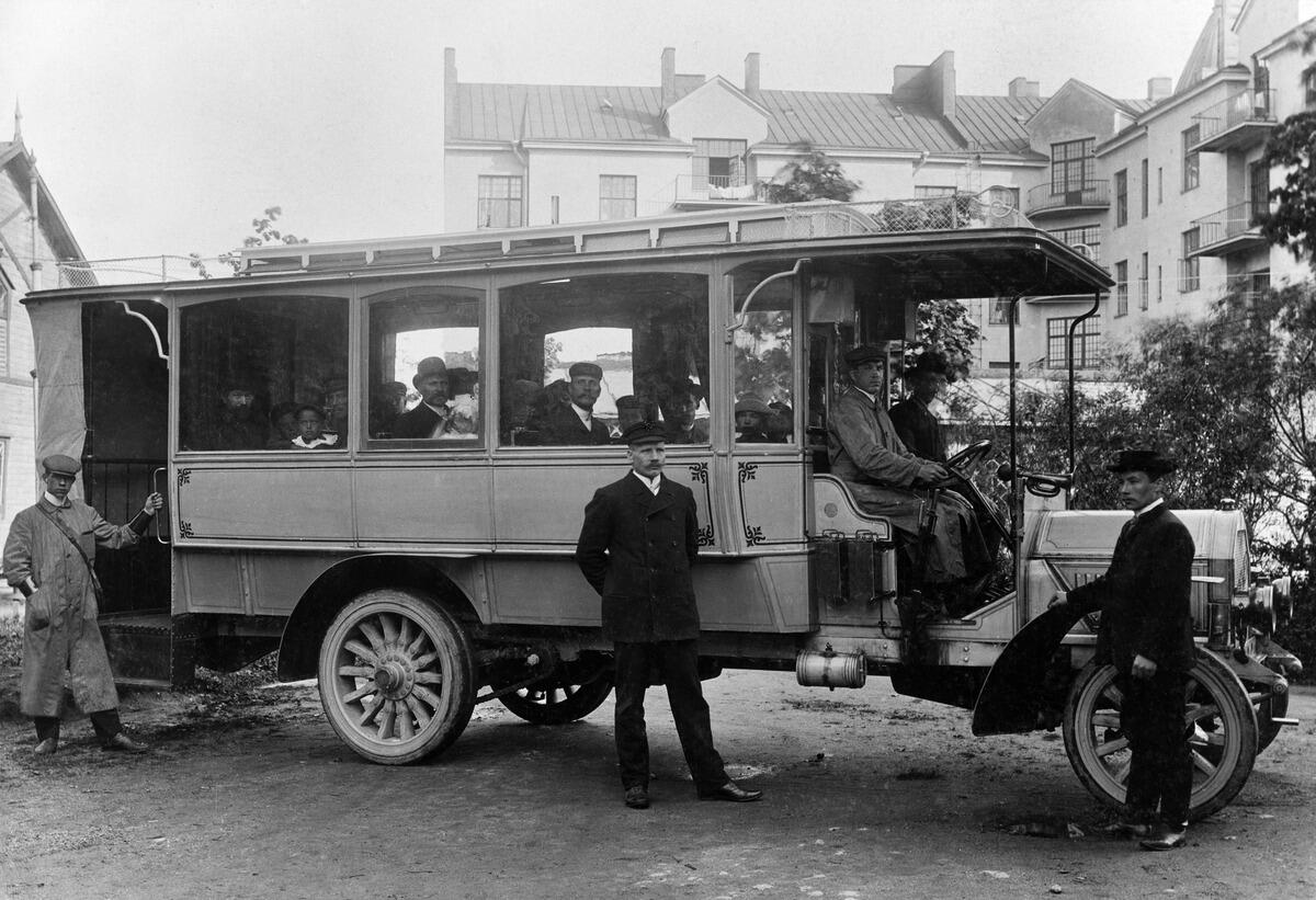 En smäcker buss med passagerare inuti. Förarsätet är separat under endast ett tak utan vindruta eller sidoväggar. Två män sitter bakom ratten. Ingången för passagerare är i bakdelen. Tre män står utanför fordonet.