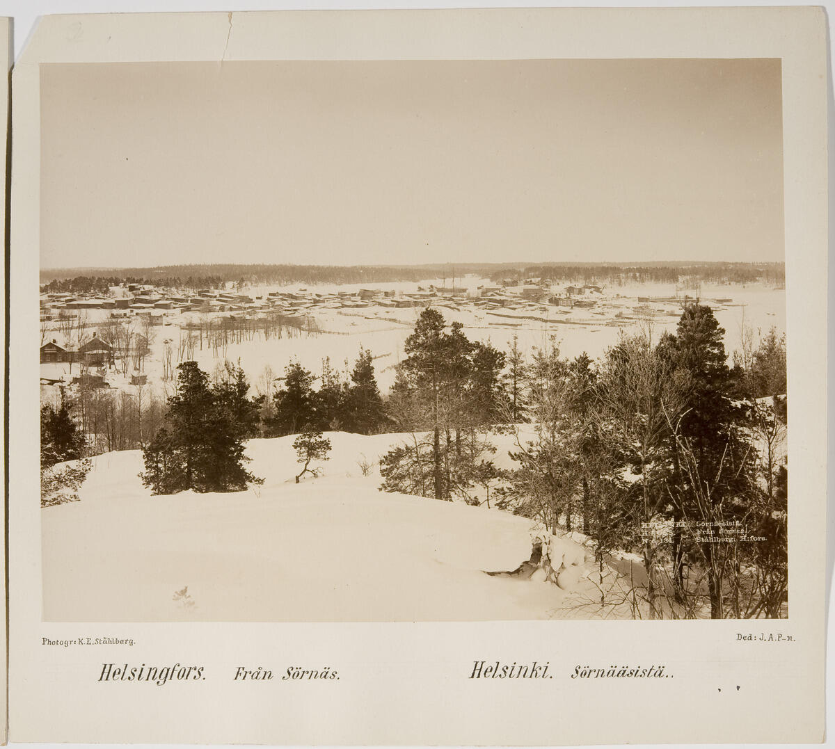 Ett snöigt landskap med träd och klippor i förgrunden, i bakgrunden låg trähusbebyggelse.