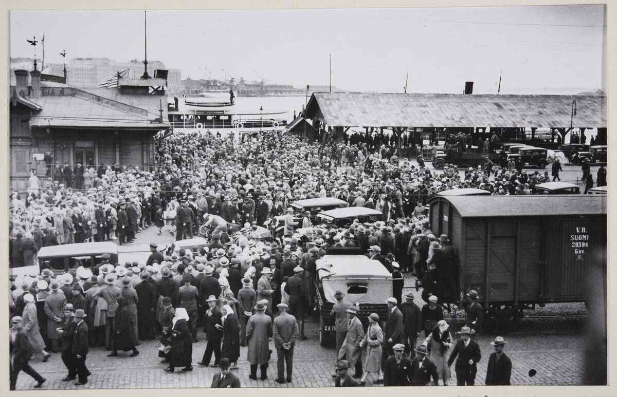 En stor folkmassa trängs i Södra Hamnen. Bland folket syns även några bilar. I bakgrunden syns hamnens låga byggnader och ett ångfartyg. Till höger i förgrunden syns en godsvagn.