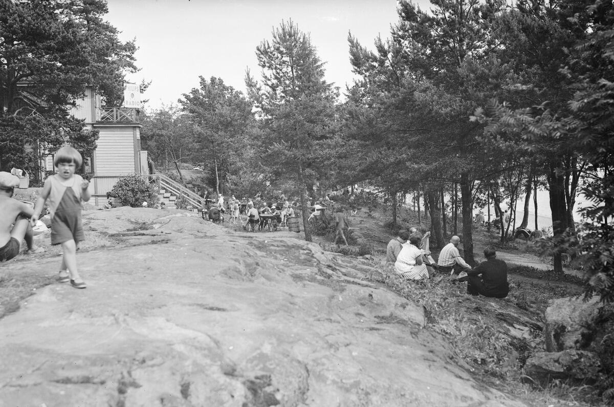 I bakgrunden till höger syns restaurangen på Blåbärslandet, till höger en strand kantad av tallar och i förgrunden klippor. Ett barn går mot fotografen, andra människor står eller sitter längre bort på restaurangens gård.