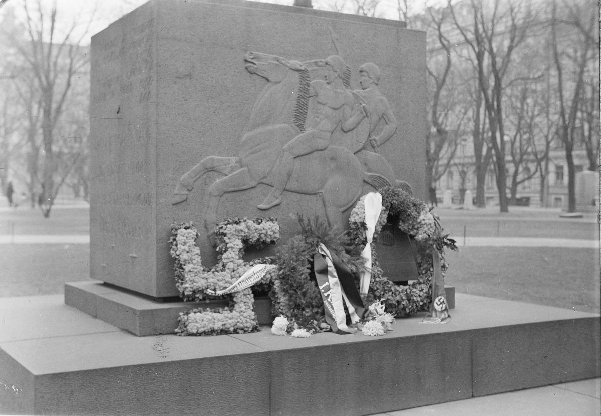 Neliskanttinen kivipaasi, johon on kuvattu kaksi hevosen selässä ratsastavaa miestä. Muistomerkin edustalle on laskettu kukkaseppeleitä.
