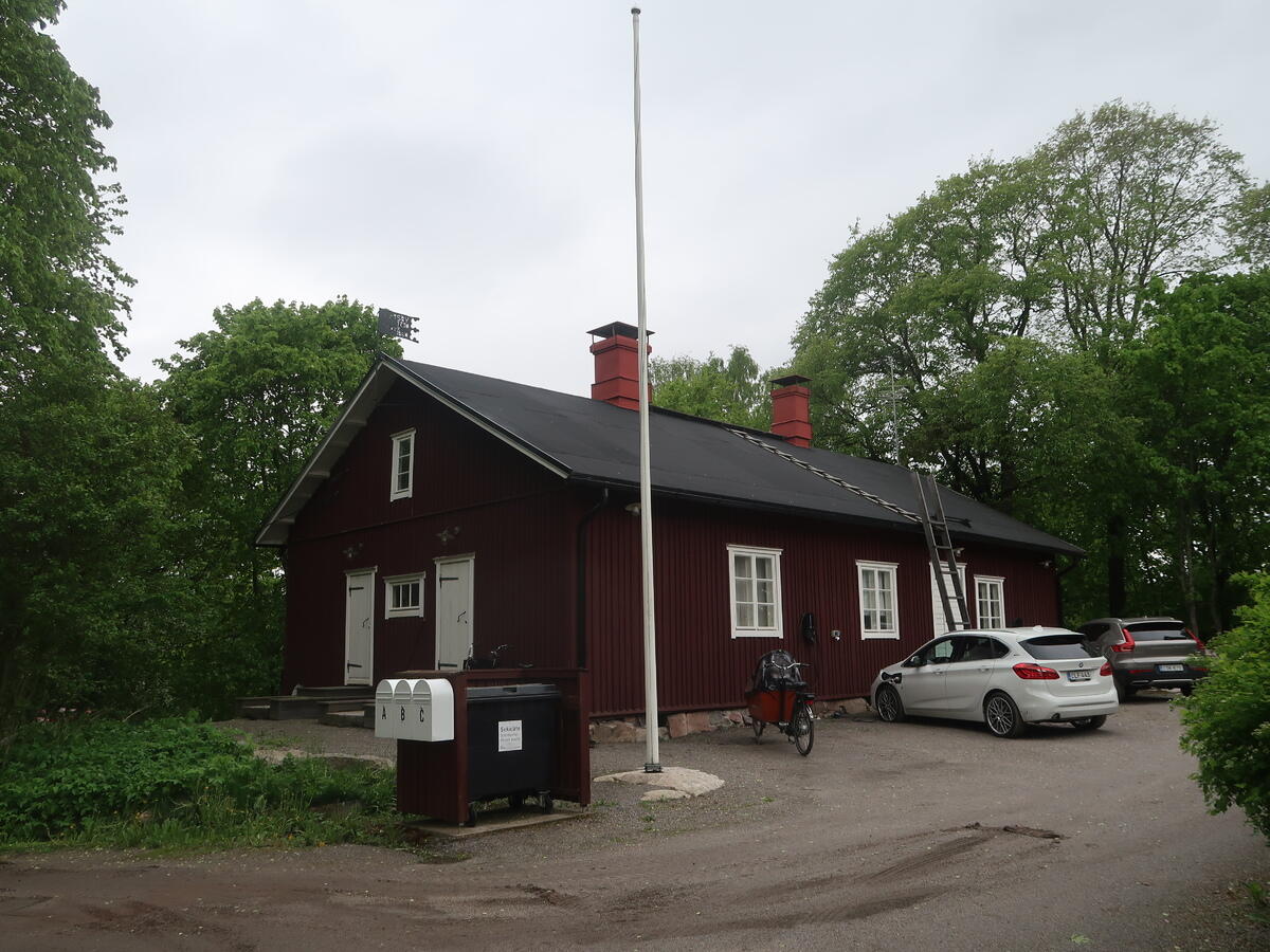 Ett mörkrött målat en och en halv vånings trähus med svart sadeltak, vita dörrar och fönsterkarmar. Fönstren är sexdelade. Framför huset står en cykel och två bilar, på gården finns en flaggstång och vid vägen står postlådor.