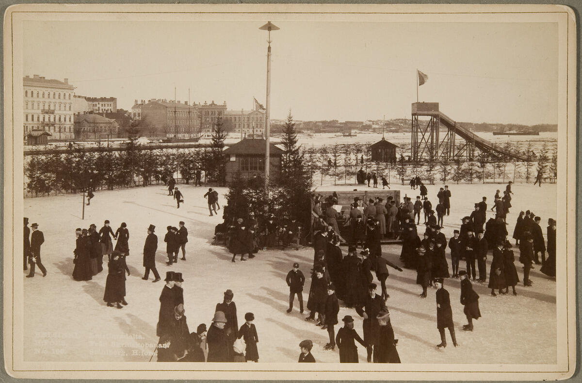 A skating rink on the ice, crowded with people. The photo also shows a wooden sledding tower and houses in Pohjoisranta.