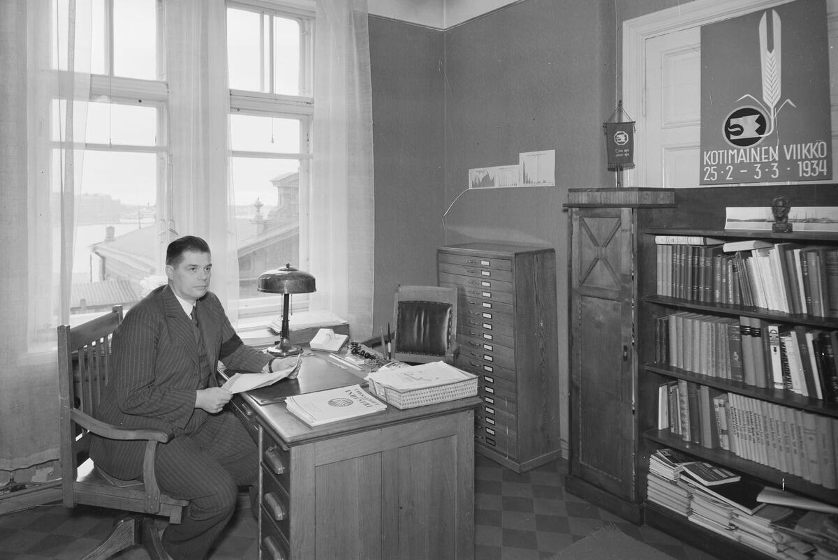 A man in a pinstripe suit sitting behind a desk with a piece of paper in his hand. There are stacks of paper and a reading lamp on the table, and a chair for a visitor on the other side of the table. The room also contains a bookshelf and a filing cabinet. You can see the sea from the window.