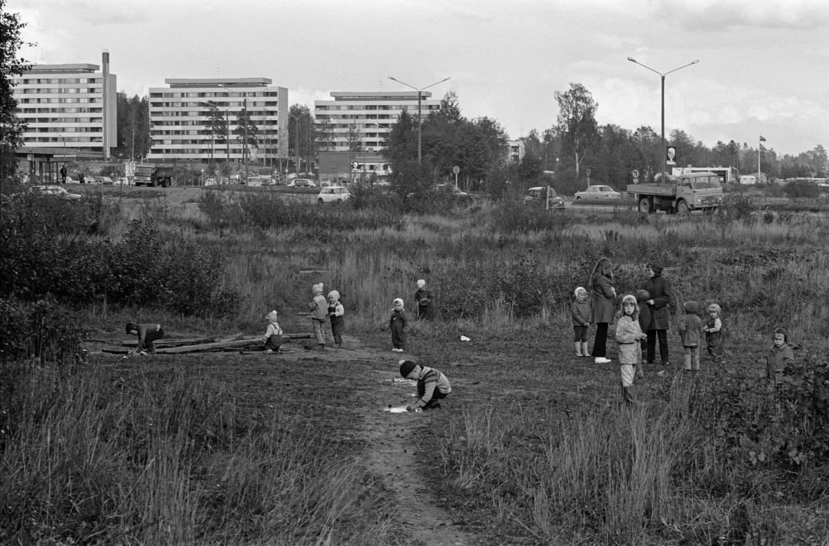Joukko lapsia leikkii joutomaa-aukealla nykyisen Itäkeskuksen kupeessa vuonna 1970. Aukean ympärillä pitkäksi kasvanutta heinikkoa ja pensaikkoa. Muutama lapsi tutkii lautakasaa, kuvan reunalla kaksi aikuista keskustelee. Taustalla Vanhanlinnankujan kerrostaloja