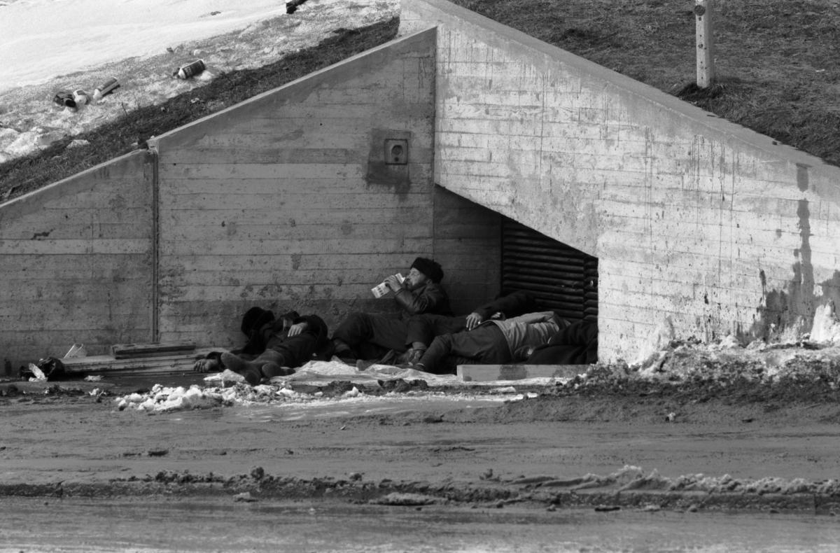 Asunnottomia miehiä makaamassa Kalliossa betonisen ilmanvaihtohormin suojassa. Toinen mies juo tölkistä piimää. Maassa on loskaa ja sen seassa voi nähdä talousspriipulloja.