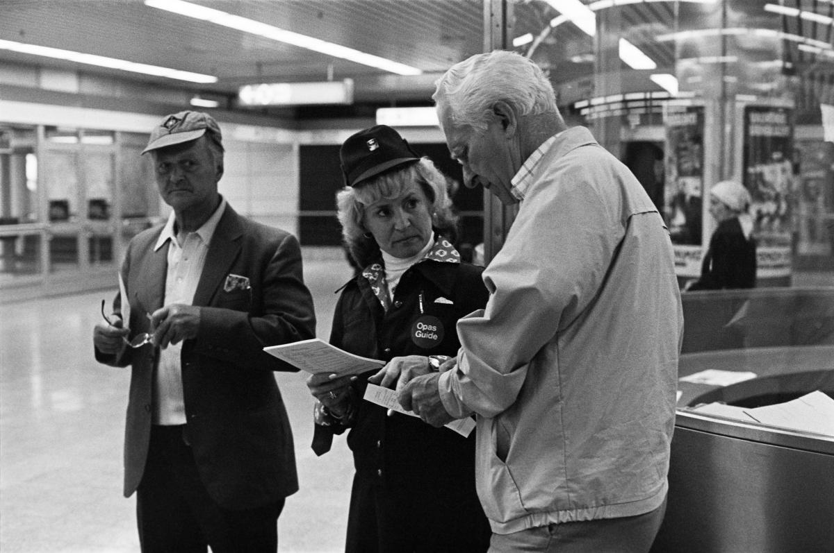 Liikennelaitoksen opas neuvomassa metron käyttäjää vuonna 1982. Virkalakkiin, takkiin ja opas-pinssiin pukeutunut nainen esittelee paperista aikataulua vanhemmalle miehelle.