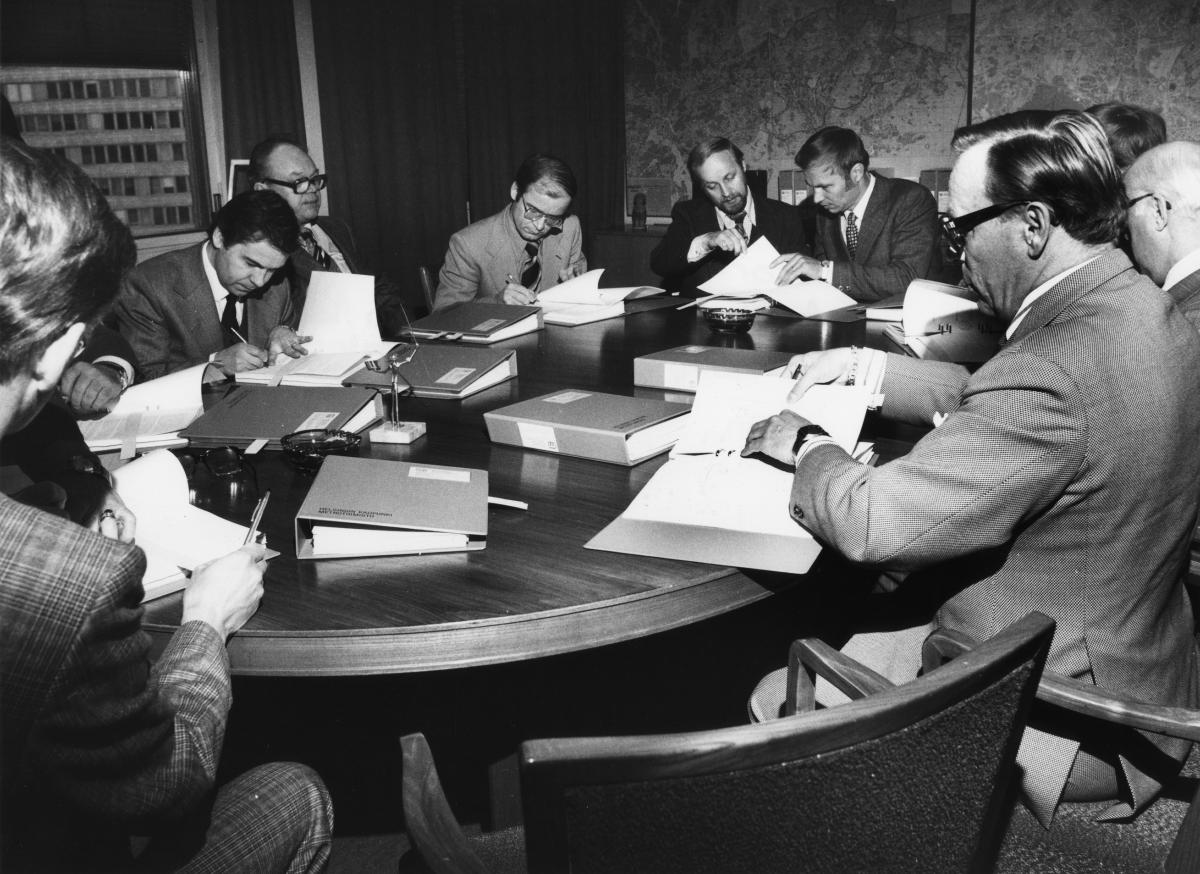 Helsingin metrotoimikunnan jäseniä kokouksessaan 1970-luvulla. Yhdeksän miestä istuu pöydän ympärillä tutkimassa asiakirjoja. Pöytä on täynnä paksuja mappeja.