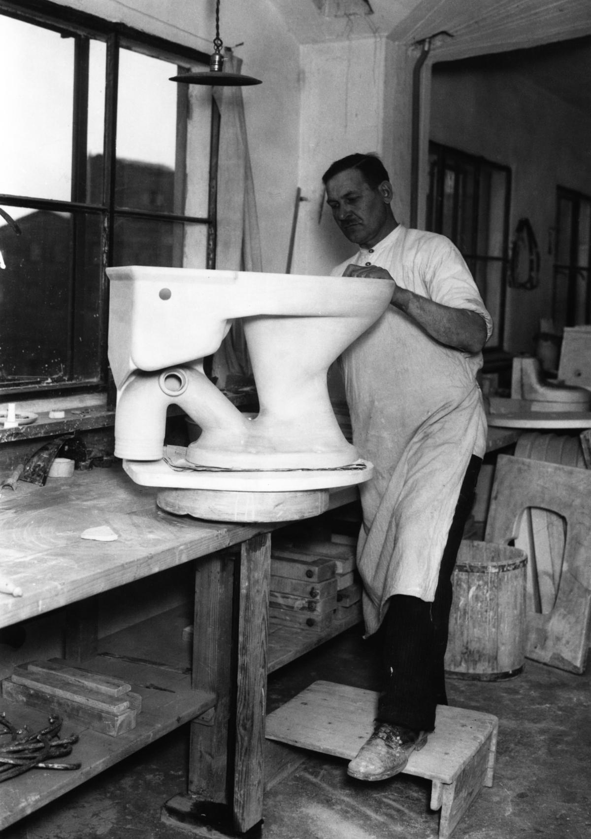 Esiliinaan pukeutunut mies muotoilee pöydän päällä käsin WC-istuinta Arabian tehtaalla.