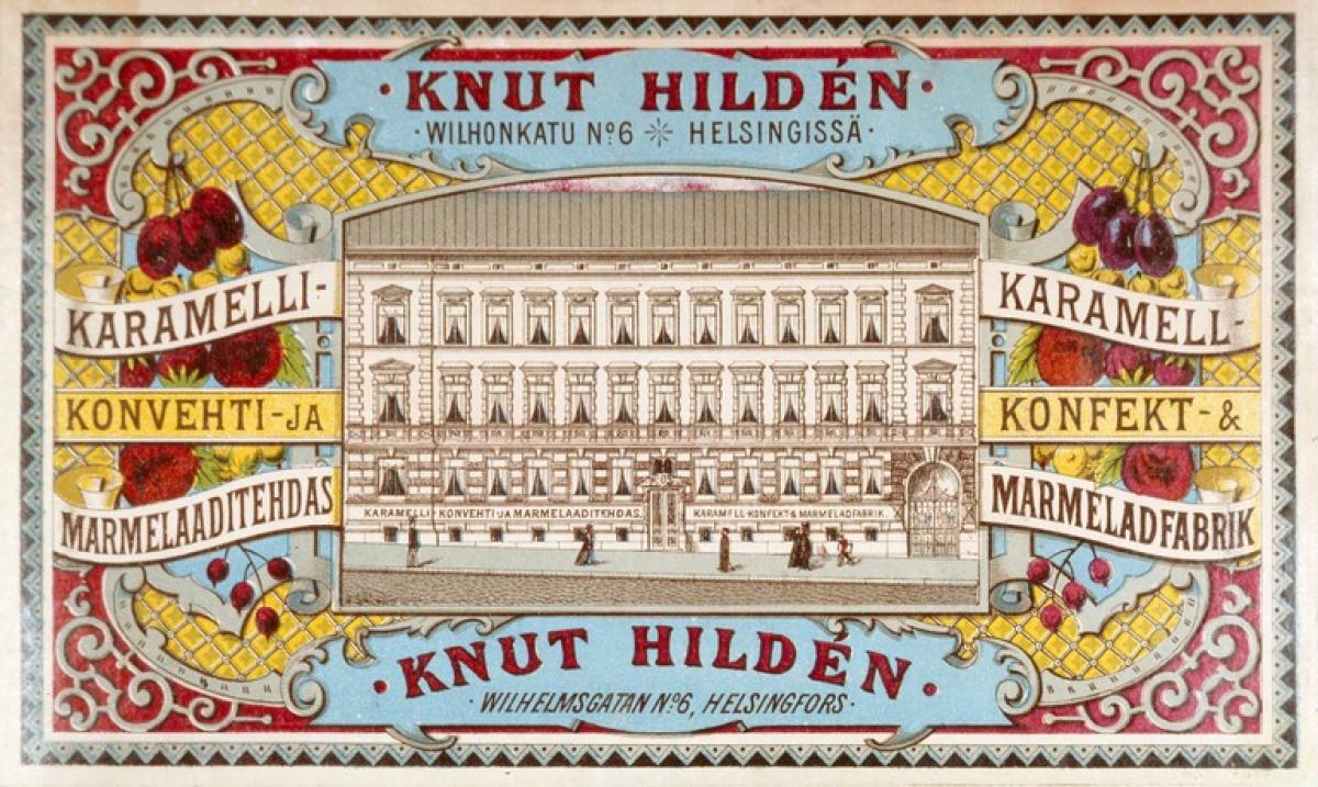 Knut Hildenin karamellitehtaan mainos, jossa keskellä tehtaan kuva. Kuvaa kehystävät hedelmäaiheiset ornamentit.