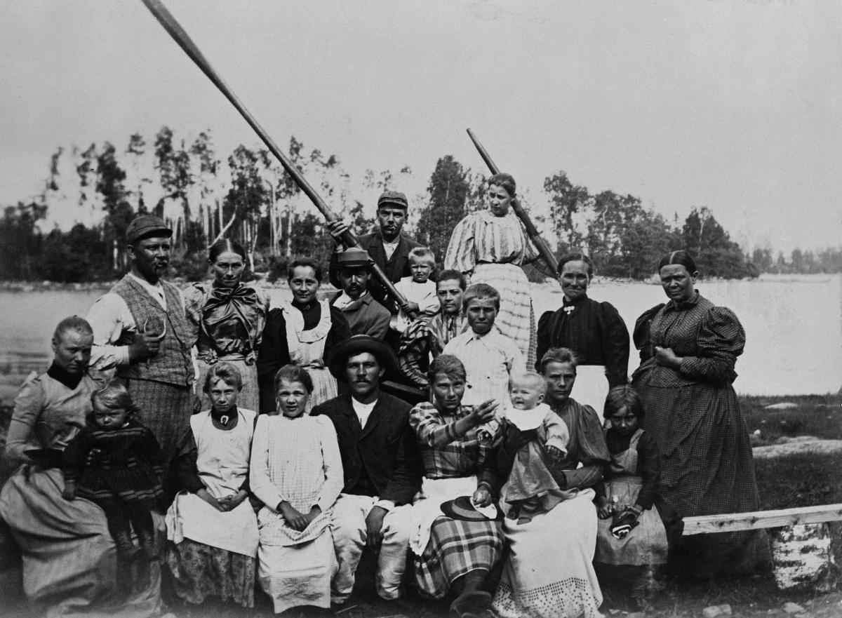 Arbetsfolk bestående av män, kvinnor och några barn sitter i en båt vid stranden.