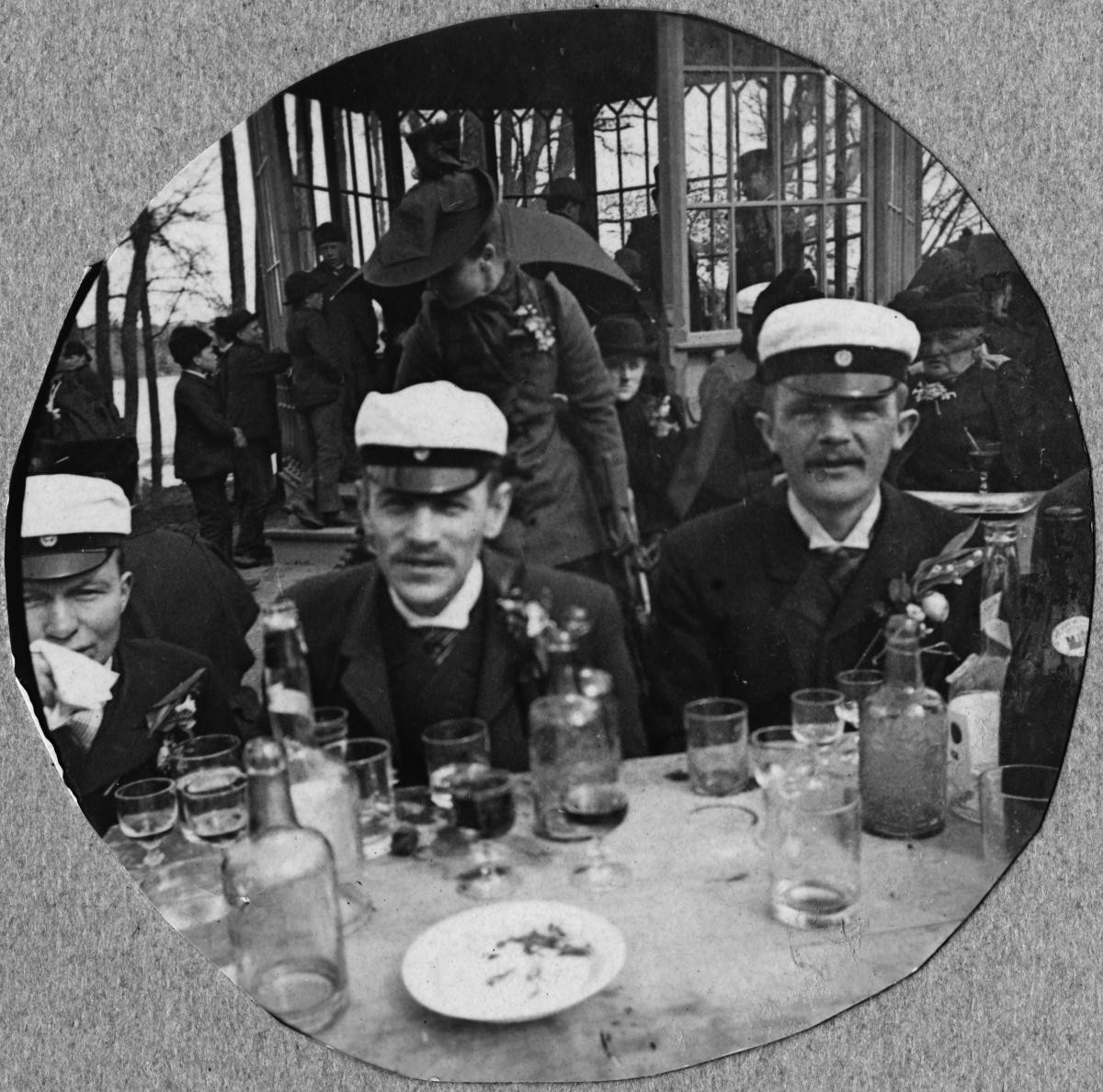 Kolme ylioppilaslakkeihin pukeutunutta miestä istuu ulkona ravintola Kaisaniemen pöydässä. Pöytä on täynnä enemmän tai vähemmän tyhjiä juomalaseja.