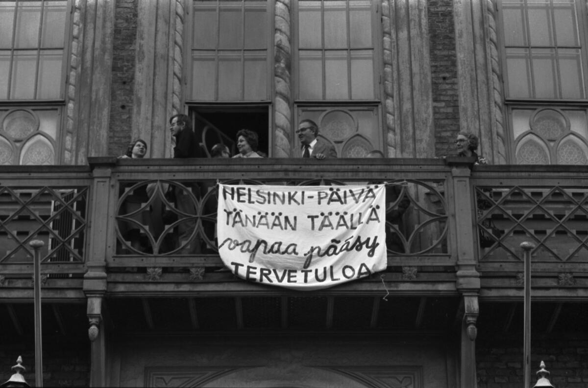 Riddarhusets balkong. Från räcket hänger ett plakat med texten Helsinki-päivä tänään täällä - vapaa pääsy - Tervetuloa