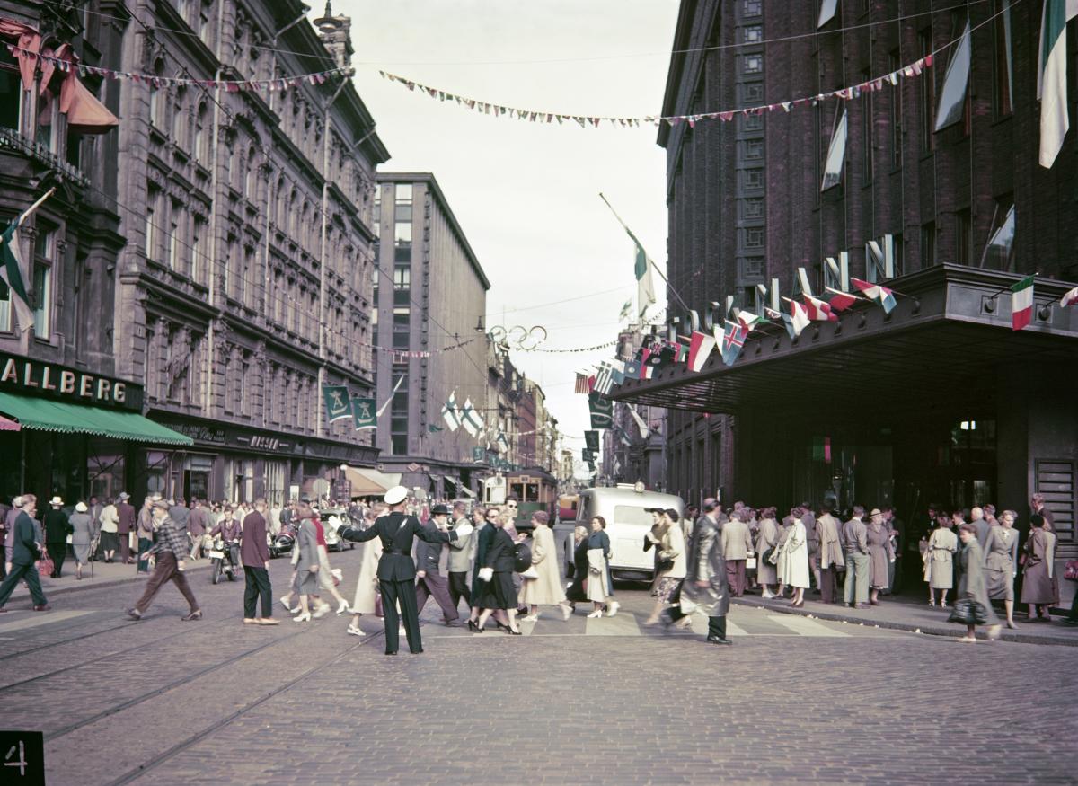 Aleksanterinkatu ja väkijoukkoa Stockmannin tavaratalon edustalta kuvattuna. Katua on koristeltu olympialaisiin osallistuvien maiden lipuilla.