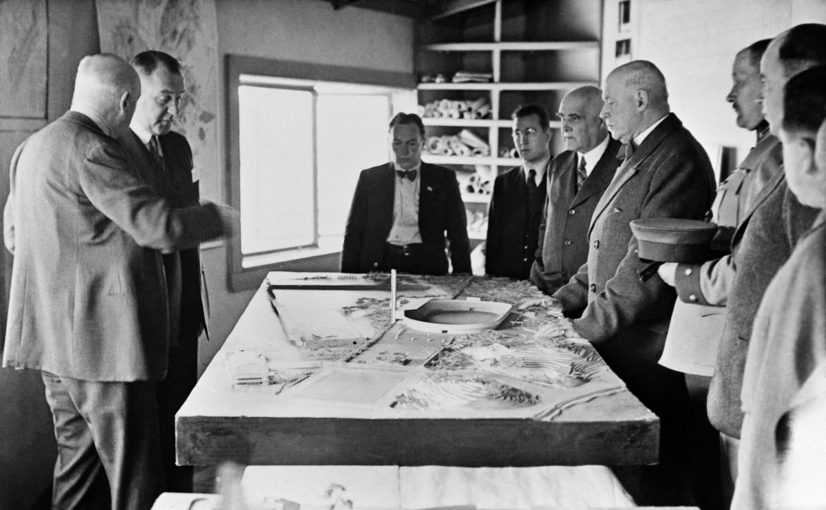 A group of men are gathered around a table, on which miniature models of Olympic sporting venues are displayed.