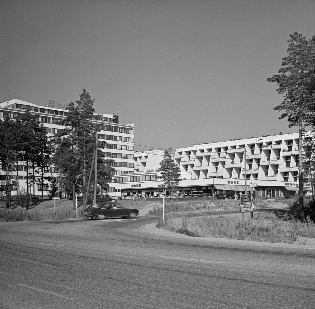Vasemmalla vaalea 1970-luvun kerrostalo, oikealla valkoinen terassitalo, jonka edessä on matala ostoskeskus. 