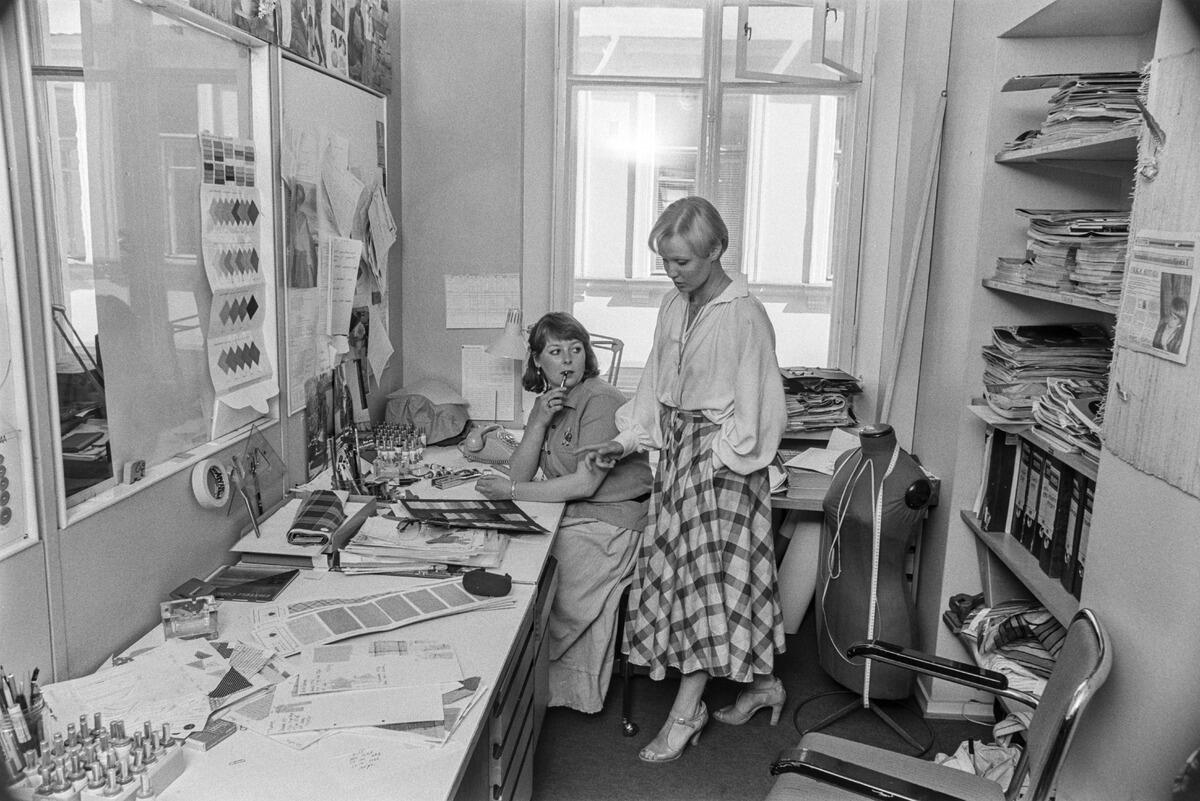 Vaatesuunnittelijat Jasmiine Julin ja Anne Linnonmaa työpöydän ääressä sivuttain katsojaan. Julin istuu ja Linnonmaa seisoo. Pöydällä on kangasnäytteitä, heidän takanaan on seinähylly, joka on täynnä julkaisuja, mappeja ja kankaita.