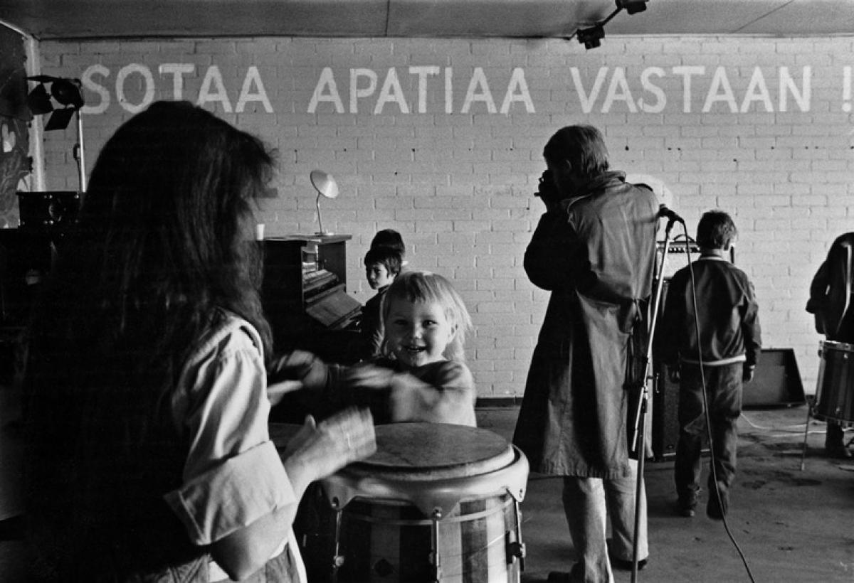 Små barn undersöker musikinstrument i Lepakkoluola. I förgrunden spelar ett litet barn stora trummor med sina händer. På väggen har målats med stora bokstäver texten ”SOTA APATIAA VASTAAN!” (Krig mot apatin!)