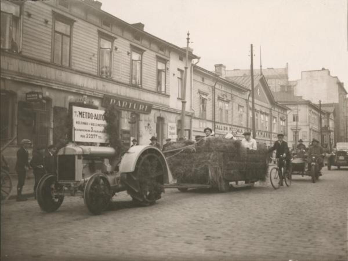 En traktor drar en vagn med hö, där människor sitter, vid Tavastvägen 12. På traktorn finns en reklam för Oy Metro-Auto, Helsingfors, Kajsaniemigatan 3, tel. 22297.