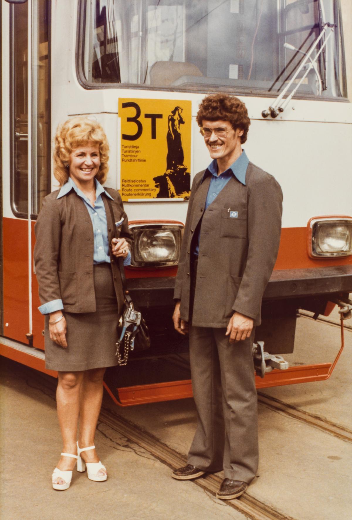 Konduktör och chaufför bär gråa uniform med ljusare skjortor. De står framför en orange spårvagn 3T. 