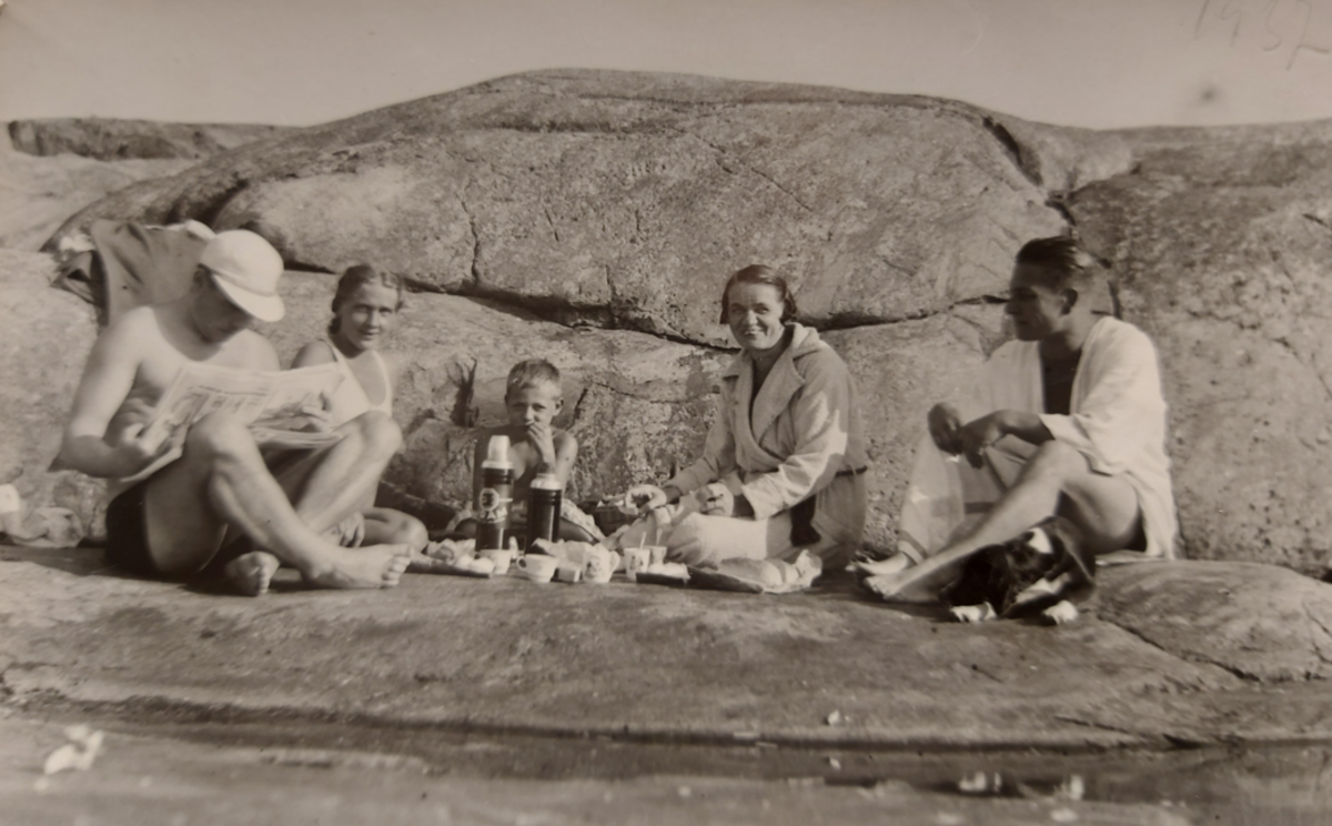 Två män och kvinnor med en pojke i mitten sitter på strandklippor med matsäck. Den ena mannen bär keps och läser en tidning. 