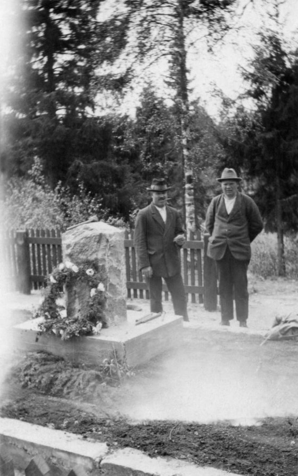 Kaksi miestä seisoo seppeleellä koristellun hautakiven vierellä.