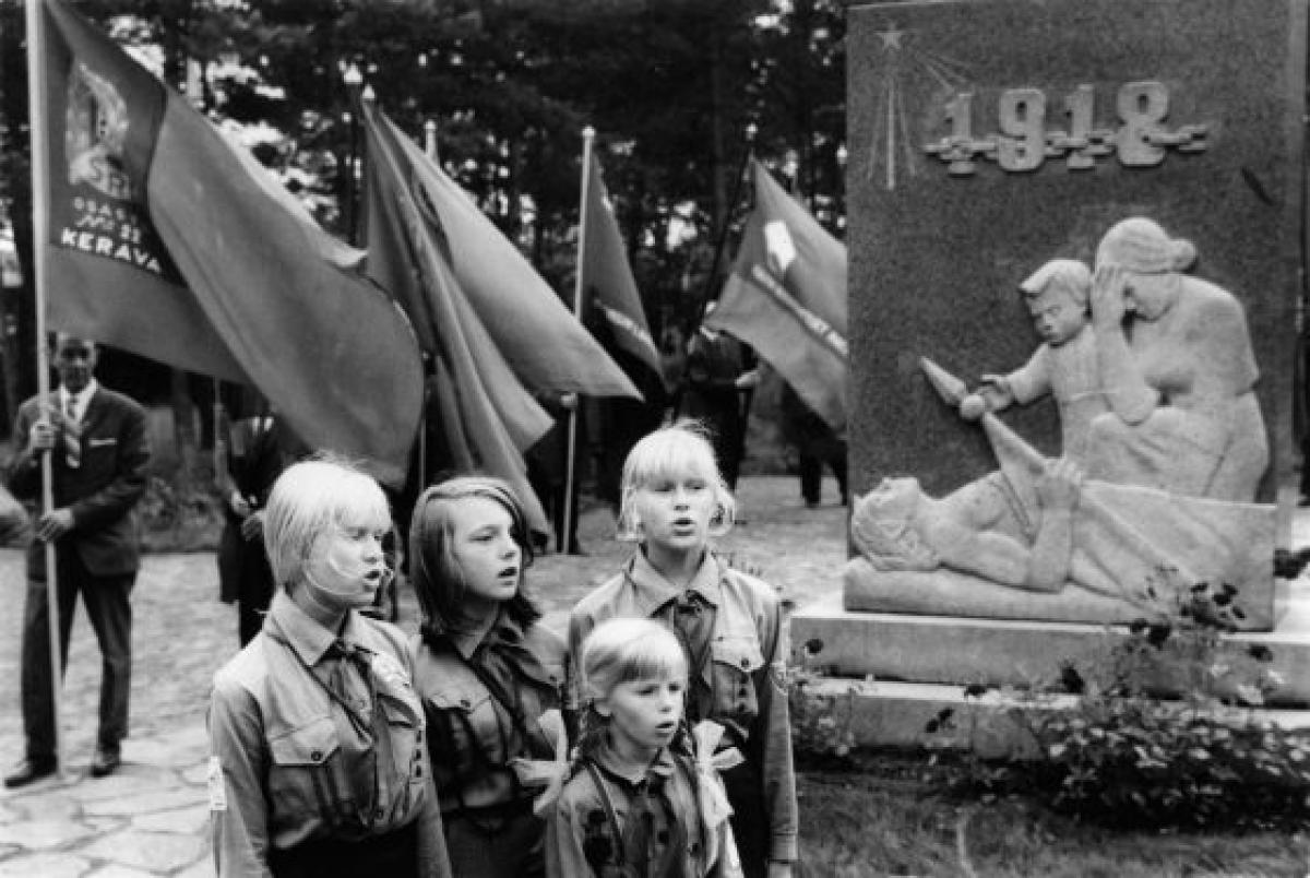 Neljä nuorta pioneeriasuista tyttöä laulaa Santahaminan veljeshaudalla punaisten muistomerkin edustalla. Taustalla lippuja piteleviä miehiä.
