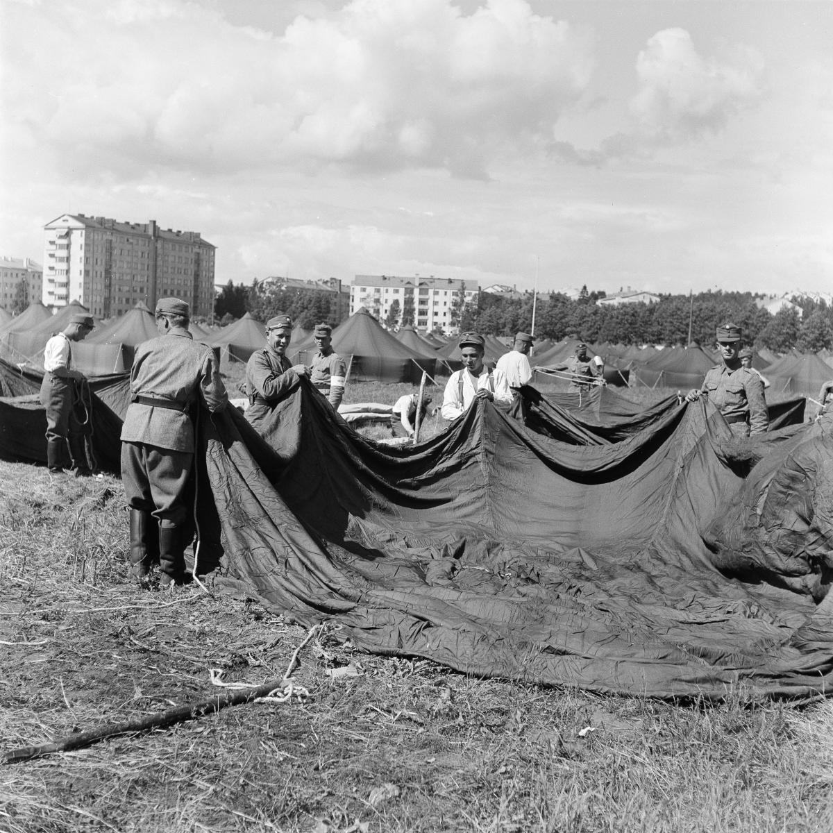 A group of soldiers is putting up a tent on a field. Block houses in the background.