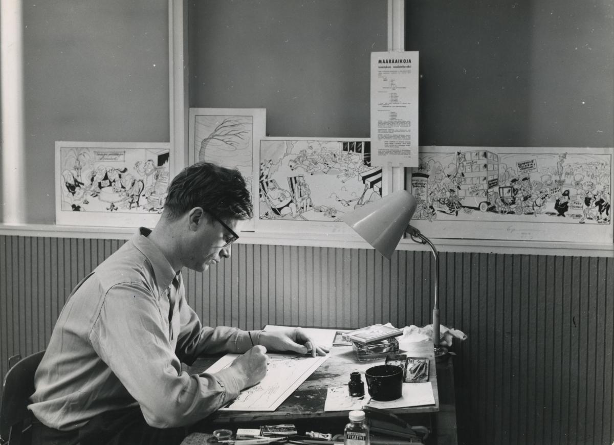 Kari Suomalainen istuu sivuttain katsojaan kirjoituspöydän ääressä piirtämässä. Hänen takanaan seinällä on monia piirroksia.