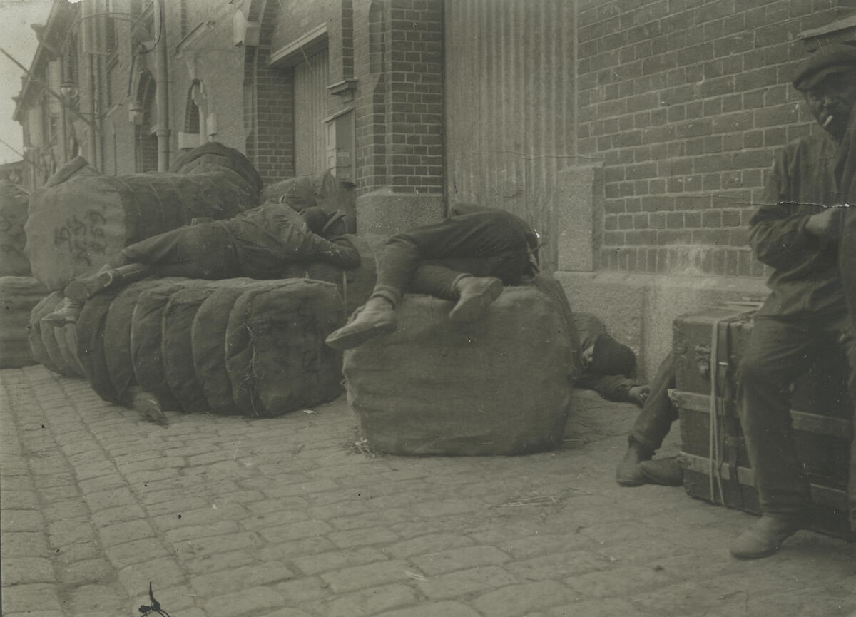 Intill magasinväggen ligger stora säckar ovanpå vilka två män ligger på sidan och vilar. En man ligger på marken bakom en säck så att endast fötterna syns. Till höger i förgrunden syns hälften av en man som sitter på en godskoffert och röker.