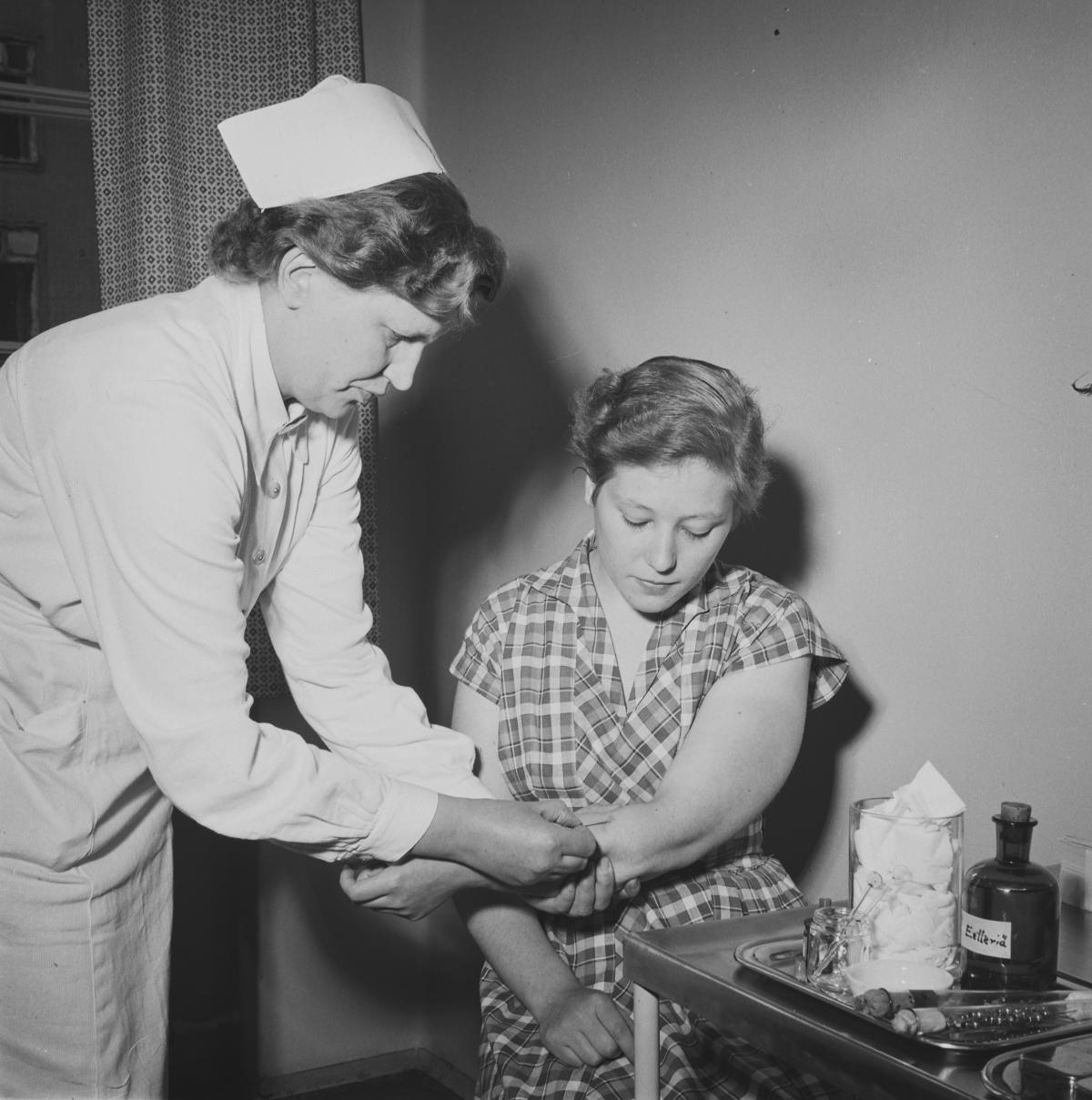 Hälsovårdare i vit hätta sticker vaccin i armen på en kvinna. På bordet bredvid kvinnan syns vaccineringstillbehör. 